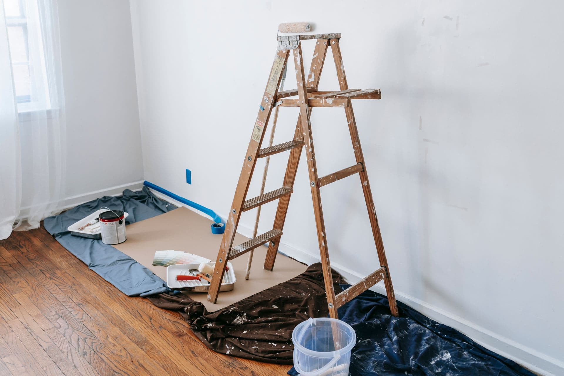 A room prepared for interior painting, with a wooden stepladder, drop cloths, paint tray, and taped baseboards