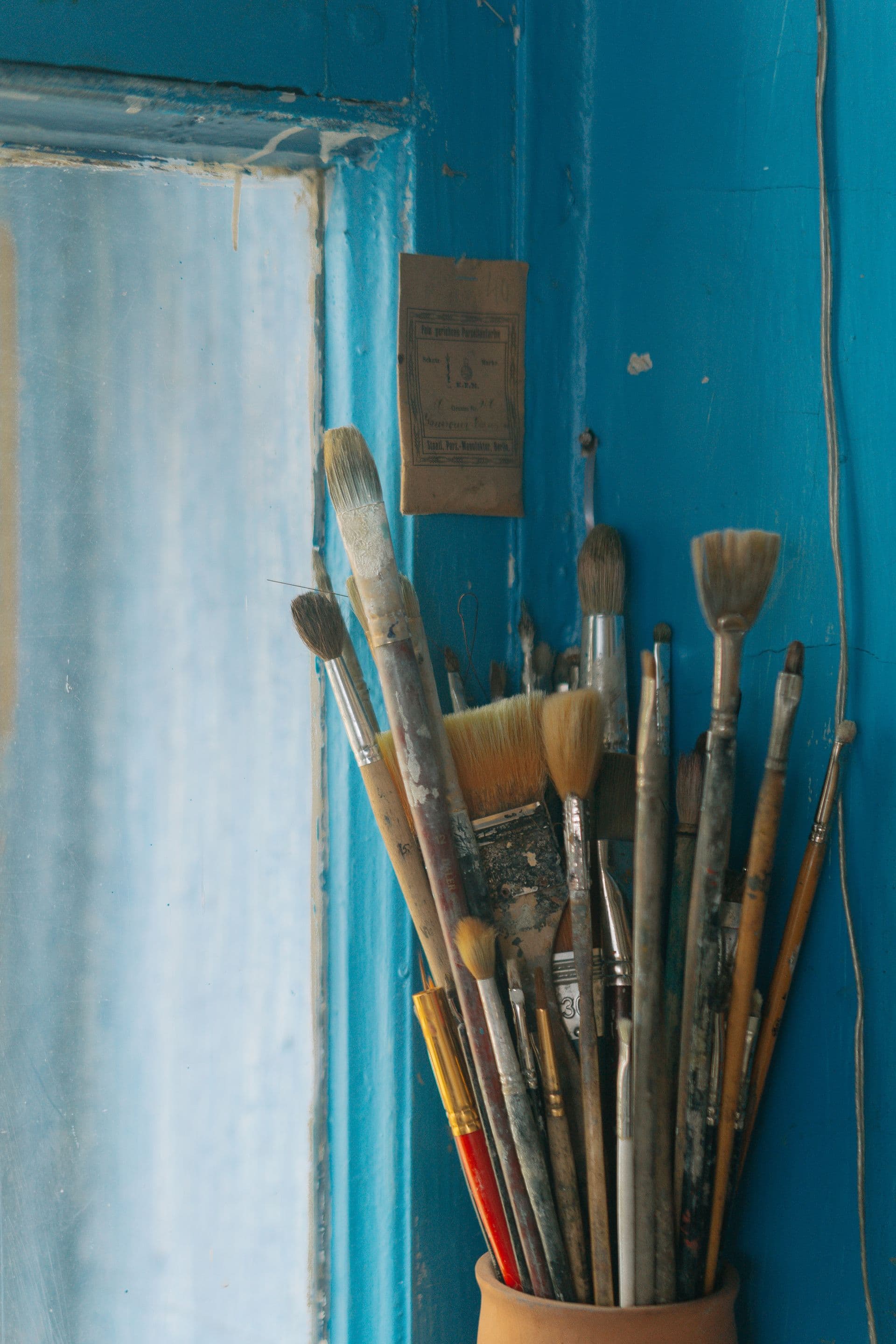 A jar of well-used paintbrushes in front of a blue-painted wall and window frame