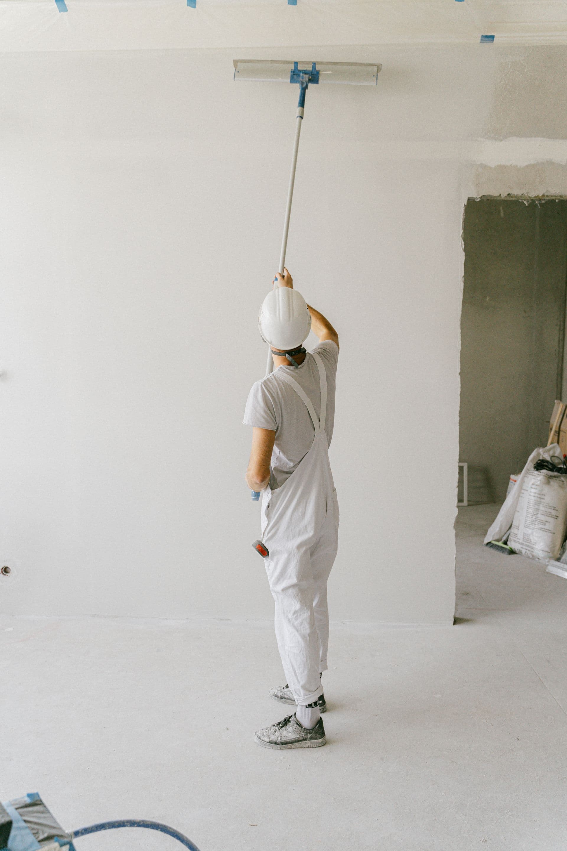 A worker in white coveralls sanding a ceiling with a pole sander before painting